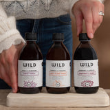 Three amber glass bottles Wild Dispensary tonics on a wooden board, with a person's hands and torso visible holding and pointing to the bottles.