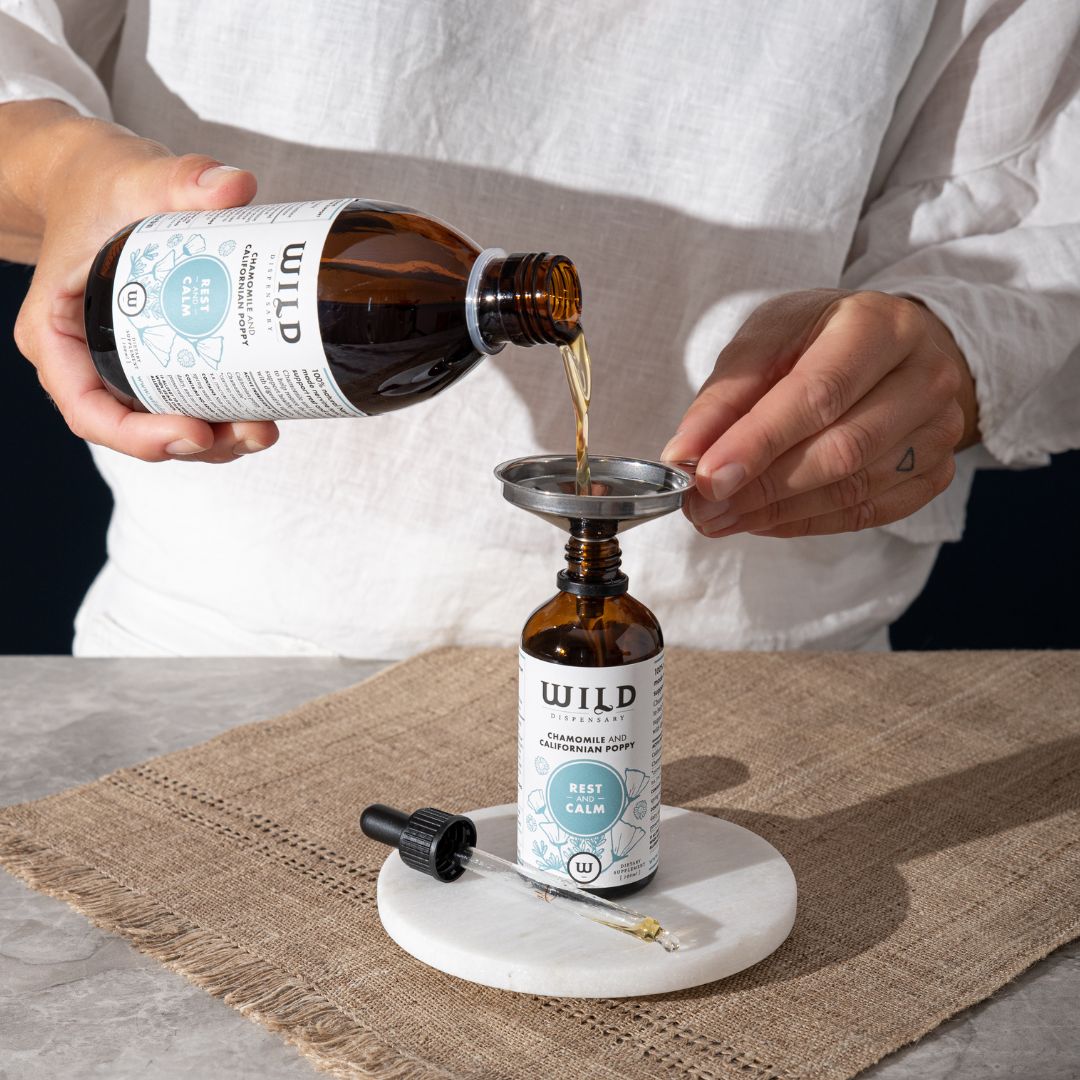 Person pouring liquid from large amber glass bottle labelled "Rest and Calm" into smaller bottle through a small metal funnel.