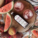 A bottle of Switchel is arranged on a round wooden board with watermelon slices, peaches, and two pink drinks with ice on a patterned blanket.