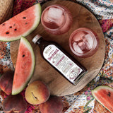 A bottle of Switchel is arranged on a round wooden board with watermelon slices, peaches, and two pink drinks with ice on a patterned blanket.