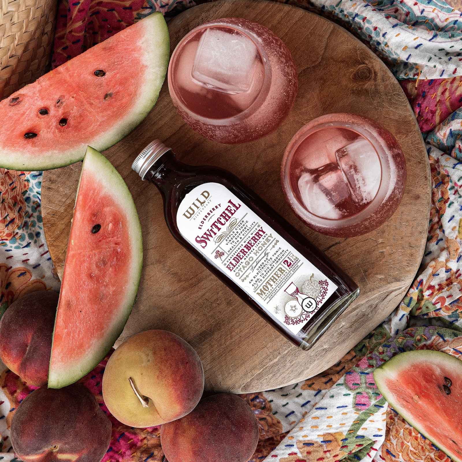 A bottle of Switchel is arranged on a round wooden board with watermelon slices, peaches, and two pink drinks with ice on a patterned blanket.