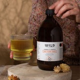 A person holds a glass of orange liquid next to a large bottle of Anti-Flam Tonic, set on a wooden surface with dried flowers against a warm neutral background.