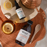 Two bottles, one labelled Anti-Flam and the other Liver Bitters, are arranged in a styled flat-lay with a copper kettle, wooden spoon, and lemon slices on a woven orange placemat.