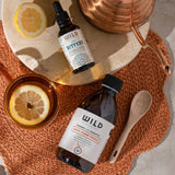 Two bottles, one labelled Anti-Flam and the other Liver Bitters, are arranged in a styled flat-lay with a copper kettle, wooden spoon, and lemon slices on a woven orange placemat.