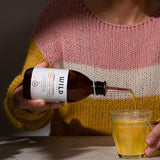 A person in a pink, cream, and mustard yellow knit sweater pours Anti-Flam into a clear glass on a tabletop.