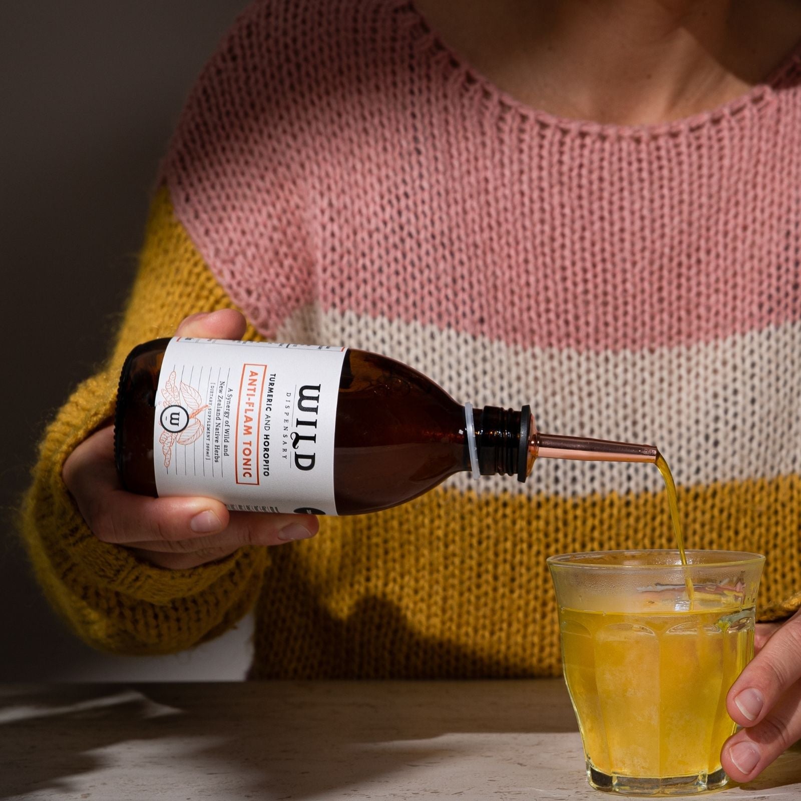 A person in a pink, cream, and mustard yellow knit sweater pours Anti-Flam into a clear glass on a tabletop.