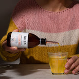 Woman in colourful jumper pouring Anti-Flam into clear glass showing orange/yellow colour of tonic.