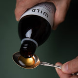 A close-up of a hand holding Chest Tonic and pouring a golden liquid into a metal spoon against a dark green background.