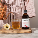 A bottle of Daily Boost Tonic stands on a wooden board beside a glass being filled with amber liquid from a copper kettle, with a person in a pink robe blurred in the background.