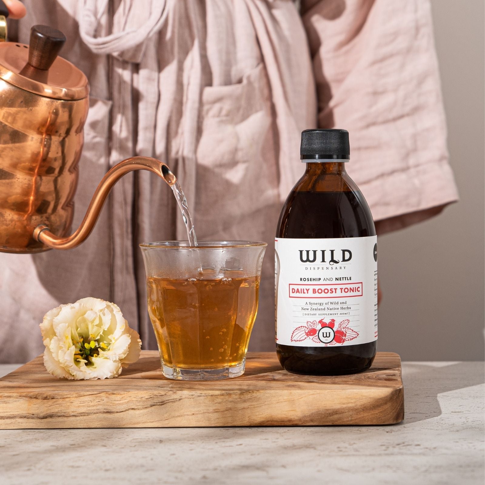 A bottle of Daily Boost Tonic stands on a wooden board beside a glass being filled with amber liquid from a copper kettle, with a person in a pink robe blurred in the background.
