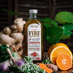 A bottle of Fire Cider surrounded by natural ingredients including fresh ginger, garlic, kawakawa leaves, and sliced oranges on a rustic wooden surface.