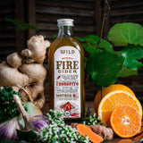 A bottle of Fire Cider surrounded by natural ingredients including fresh ginger, garlic, kawakawa leaves, and sliced oranges on a rustic wooden surface.