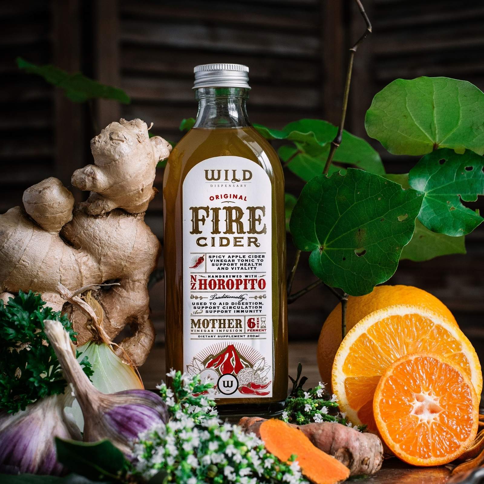 A bottle of Fire Cider surrounded by natural ingredients including fresh ginger, garlic, kawakawa leaves, and sliced oranges on a rustic wooden surface.
