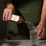 Person pouring Fire Cider into a clear glass with water using the tonic pourer which fits into the lid of the bottle.