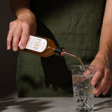 Person pouring Fire Cider into a clear glass with water using the tonic pourer which fits into the lid of the bottle.