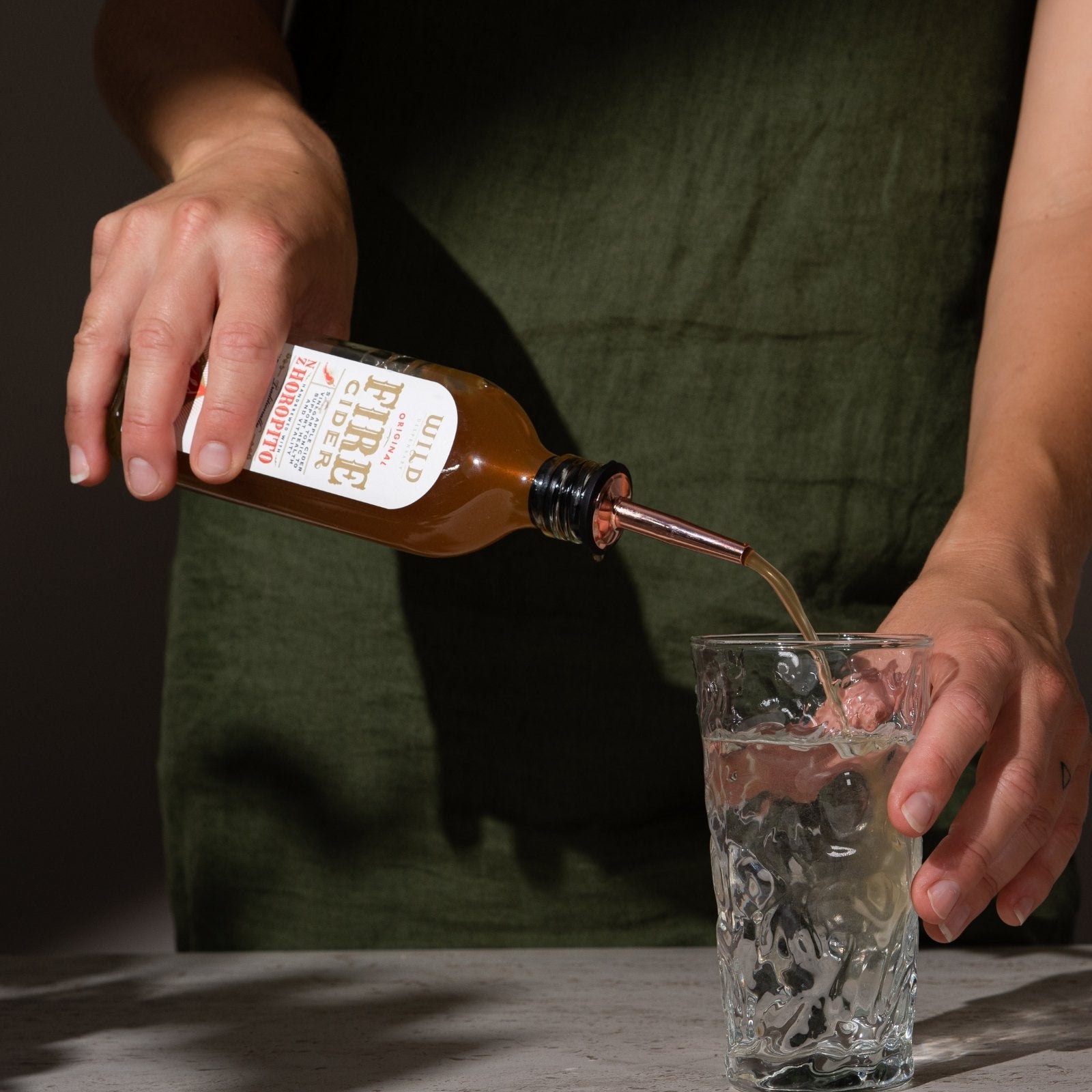 Person pouring Fire Cider into a clear glass with water using the tonic pourer which fits into the lid of the bottle.