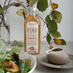 A bottle of Fire Cider stands on a light grey tabletop styled with a salad bowl, a ceramic pitcher on a saucer, and autumn leaves in a glass vase against a neutral background.