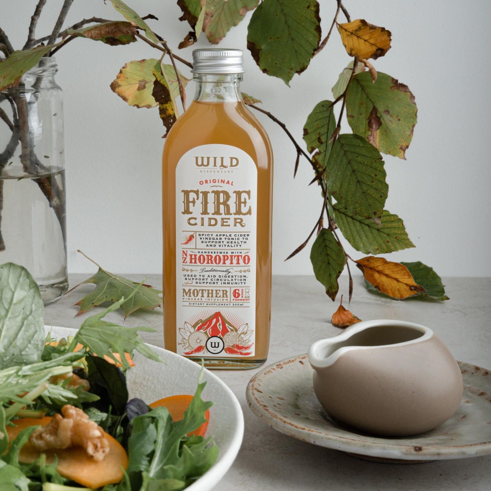 A bottle of Fire Cider stands on a light grey tabletop styled with a salad bowl, a ceramic pitcher on a saucer, and autumn leaves in a glass vase against a neutral background.