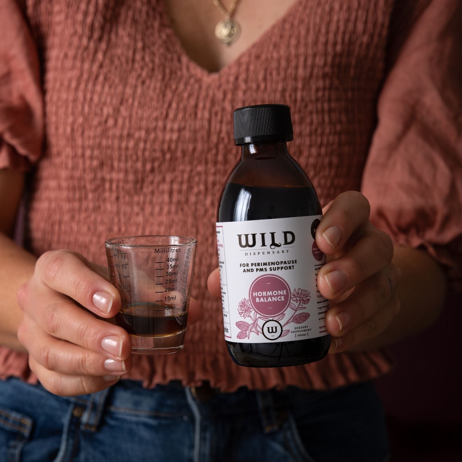 A person in a rust-colored top holds a dark amber bottle labelled Hormone Balance and a measuring cup containing the liquid.