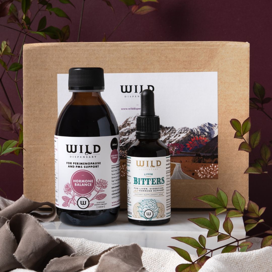 A gift pack from Wild Dispensary featuring two bottles, one labelled 'Hormone Balance' and the other 'Liver Bitters', presented in front of a box. The products are displayed on a fabric surface with leaves around them.