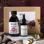 A gift pack from Wild Dispensary featuring two bottles, one labelled 'Hormone Balance' and the other 'Liver Bitters', presented in front of a box. The products are displayed on a fabric surface with leaves around them.