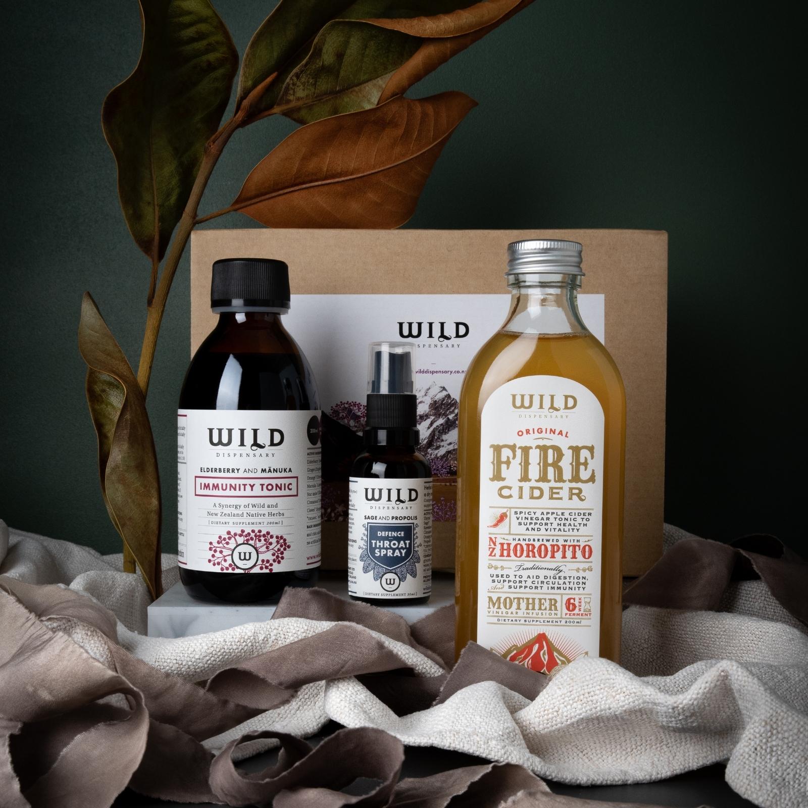 A curated gift set including an Immunity Tonic, Fire Cider and Throat Spray arranged on draped fabric in front of a gift box with decorative leaves.