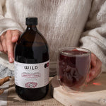 A person in a cream knit sweater holds a dark amber bottle labelled Immunity Tonic next to a clear glass filled with the dark red liquid, set on a wooden board with dried botanicals.