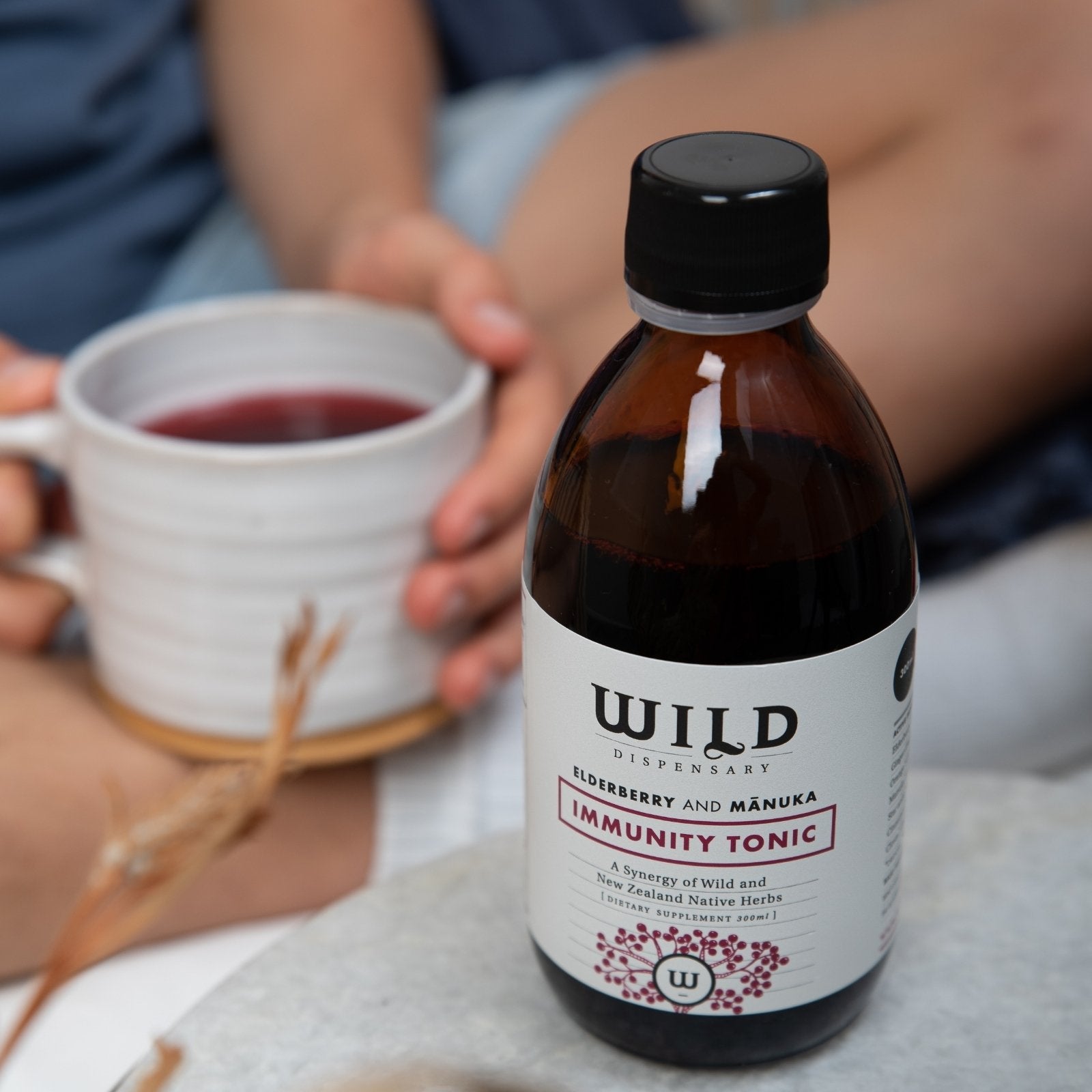An amber glass bottle of Immunity Tonic with a black cap and white label, set on a table with a person holding a white mug in the blurred background.