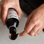 A close-up of a person's hands holding a dark glass bottle labelled Immunity Tonic and pouring a dark red liquid into a metal spoon over a light grey surface.