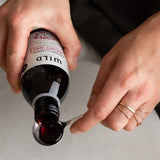 A close-up of a person's hands holding a dark glass bottle labelled Immunity Tonic and pouring a dark red liquid into a metal spoon over a light grey surface.