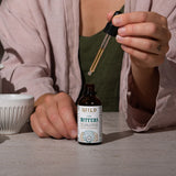 A person holds a glass dropper above an amber bottle of Liver Bitters on a tabletop, with a white cup partially visible at left.