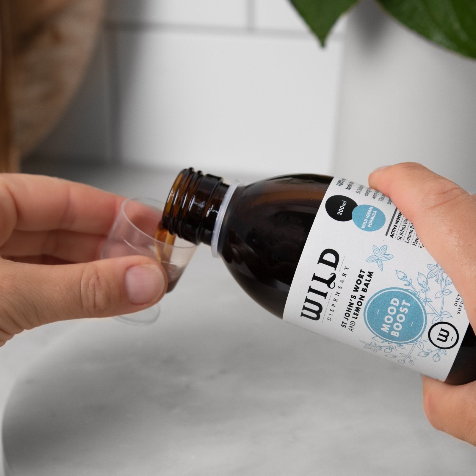 A person pours a dark amber liquid from a bottle labelled Mood Boost into a small clear measuring cup in a kitchen setting.