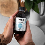 A person's hands holding a bottle of Mood Boost tincture against a soft-focus background of a white vase with green leaves.