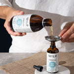 A person in a white shirt pours Rest and Calm bulk into a smaller amber dropper bottle using a metal funnel. 