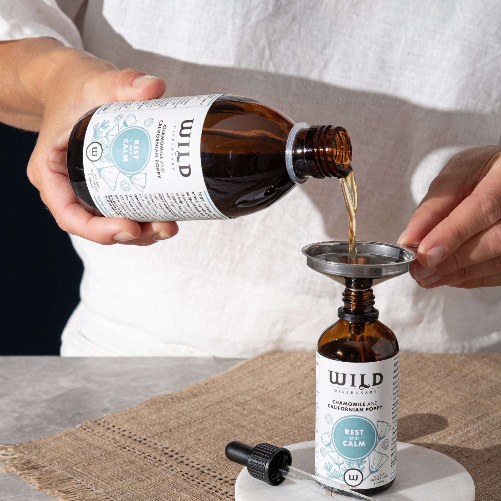 A person in a white shirt pours Rest and Calm bulk into a smaller amber dropper bottle using a metal funnel. 