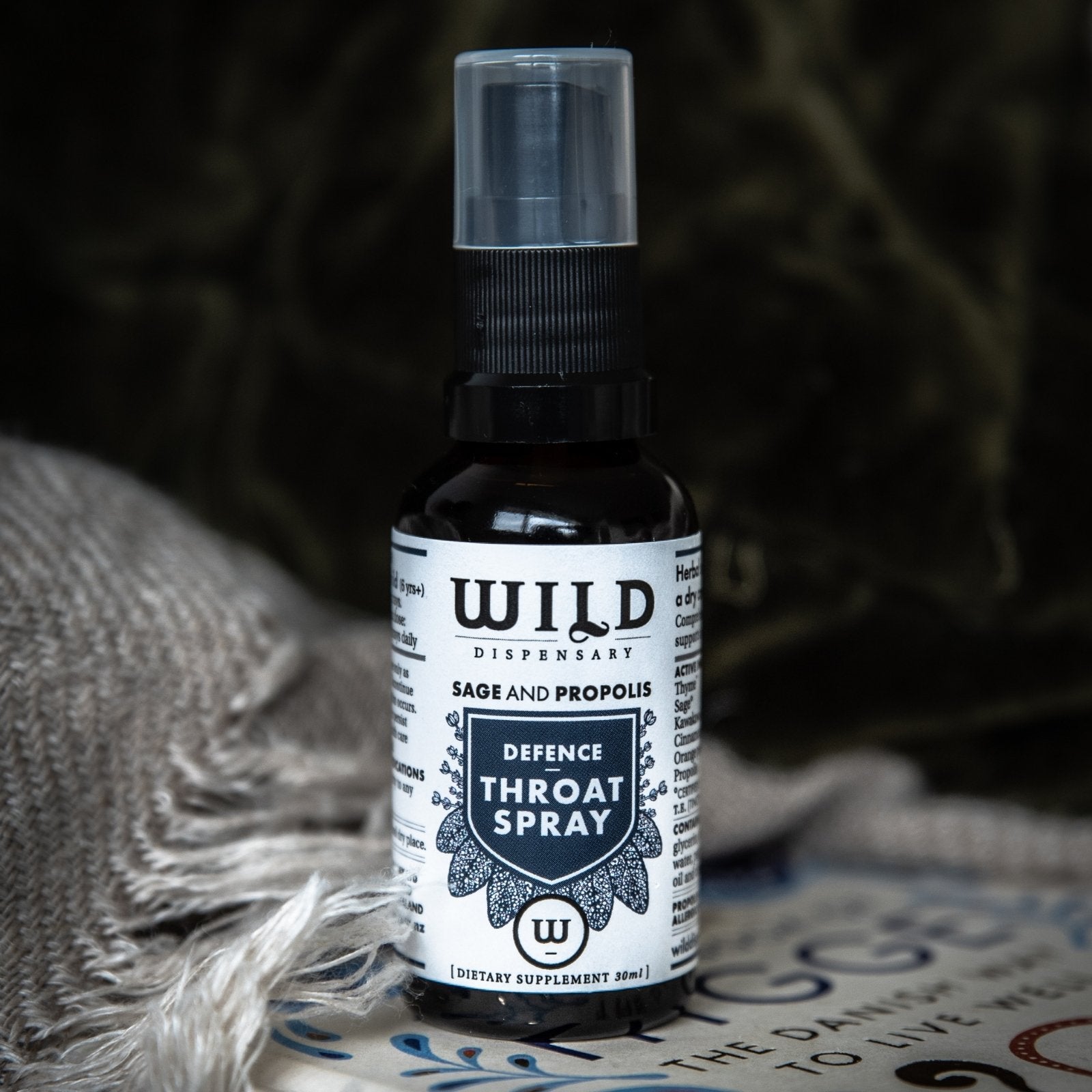 A bottle of Wild Dispensary Defence Throat Spray shown on a patterned surface with a textured grey blanket in the foreground and a dark, atmospheric background.