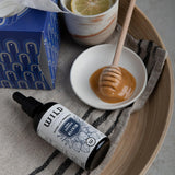 A bottle of Vira Defence Elixir shown next to a white bowl of honey with a wooden dipper, a blue tissue box, and a ceramic cup with a lemon slice, all arranged on a striped cloth inside a wooden tray.